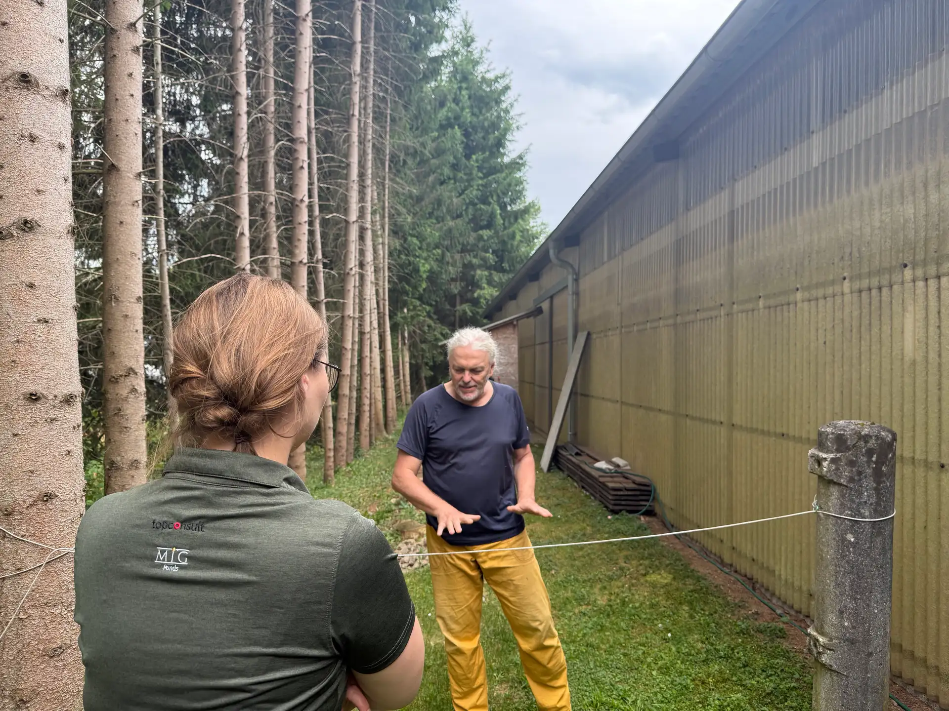 Trainer Albert Iglseder im direkten Austausch – praxisnah und auf Augenhöhe. Mann in gelbem Shirt erklärt einer Frau eine Übung mit einem gespannten Seil zwischen Bäumen und einer Wand.