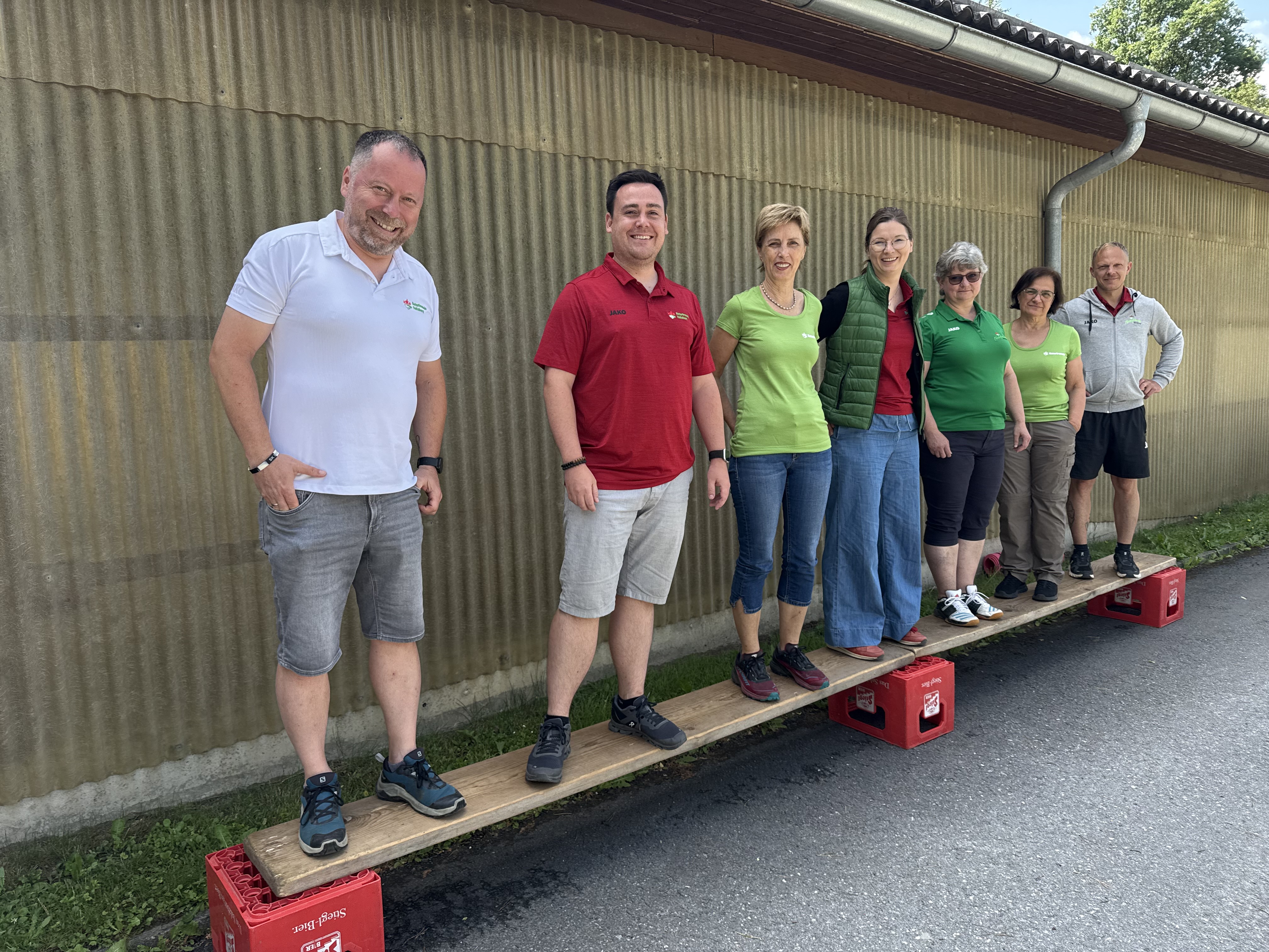 Gemeinsam die Balance halten – buchstäblich. Die Teamübung auf dem Balancebrett war ein Highlight des Wochenendes. Sieben Personen stehen nebeneinander auf einem schmalen Holzbrett, das auf roten Bierkisten liegt, und lachen in die Kamera.