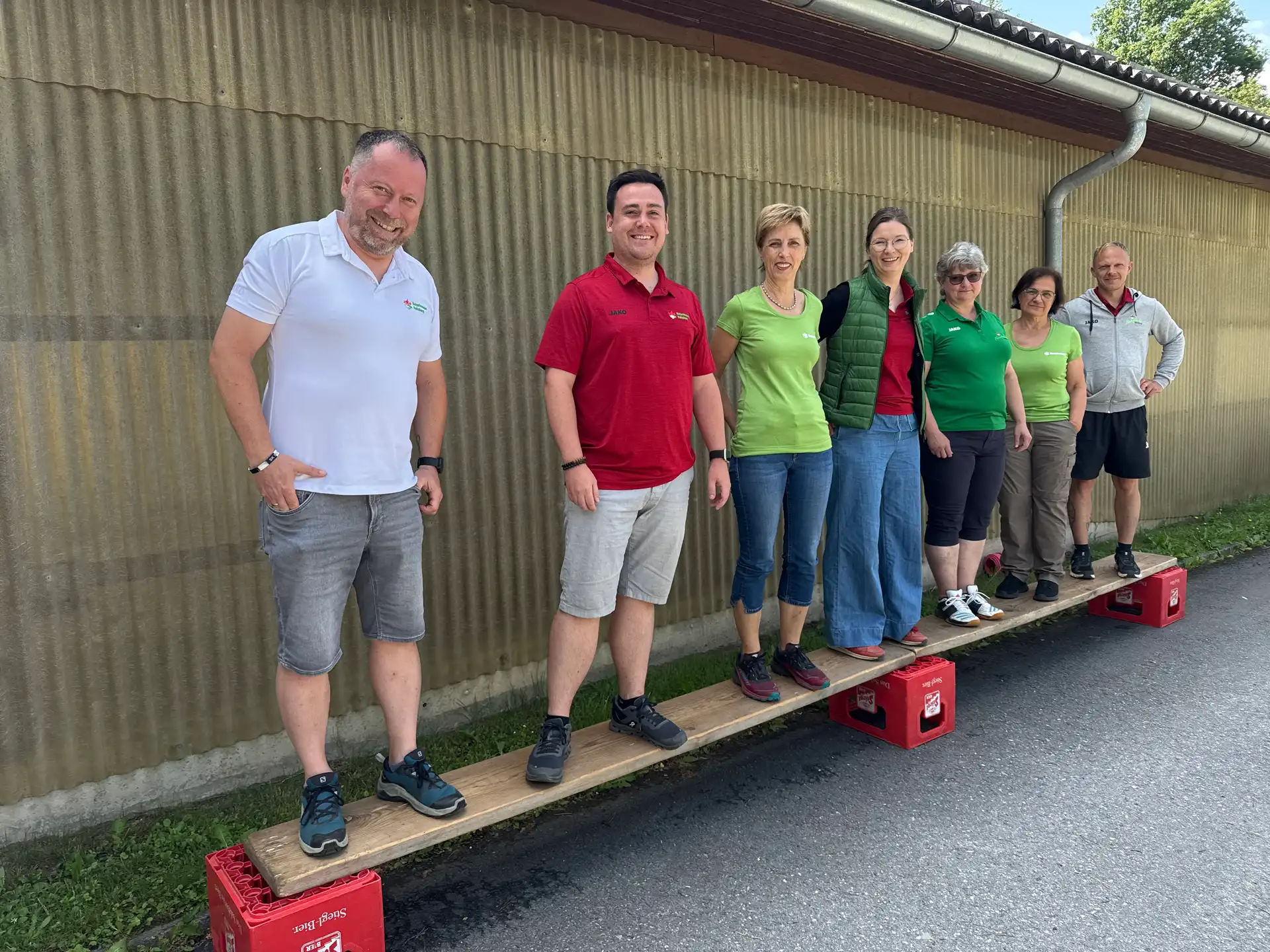 Gemeinsam die Balance halten – buchstäblich. Die Teamübung auf dem Balancebrett war ein Highlight des Wochenendes. Sieben Personen stehen nebeneinander auf einem schmalen Holzbrett, das auf roten Bierkisten liegt, und lachen in die Kamera.