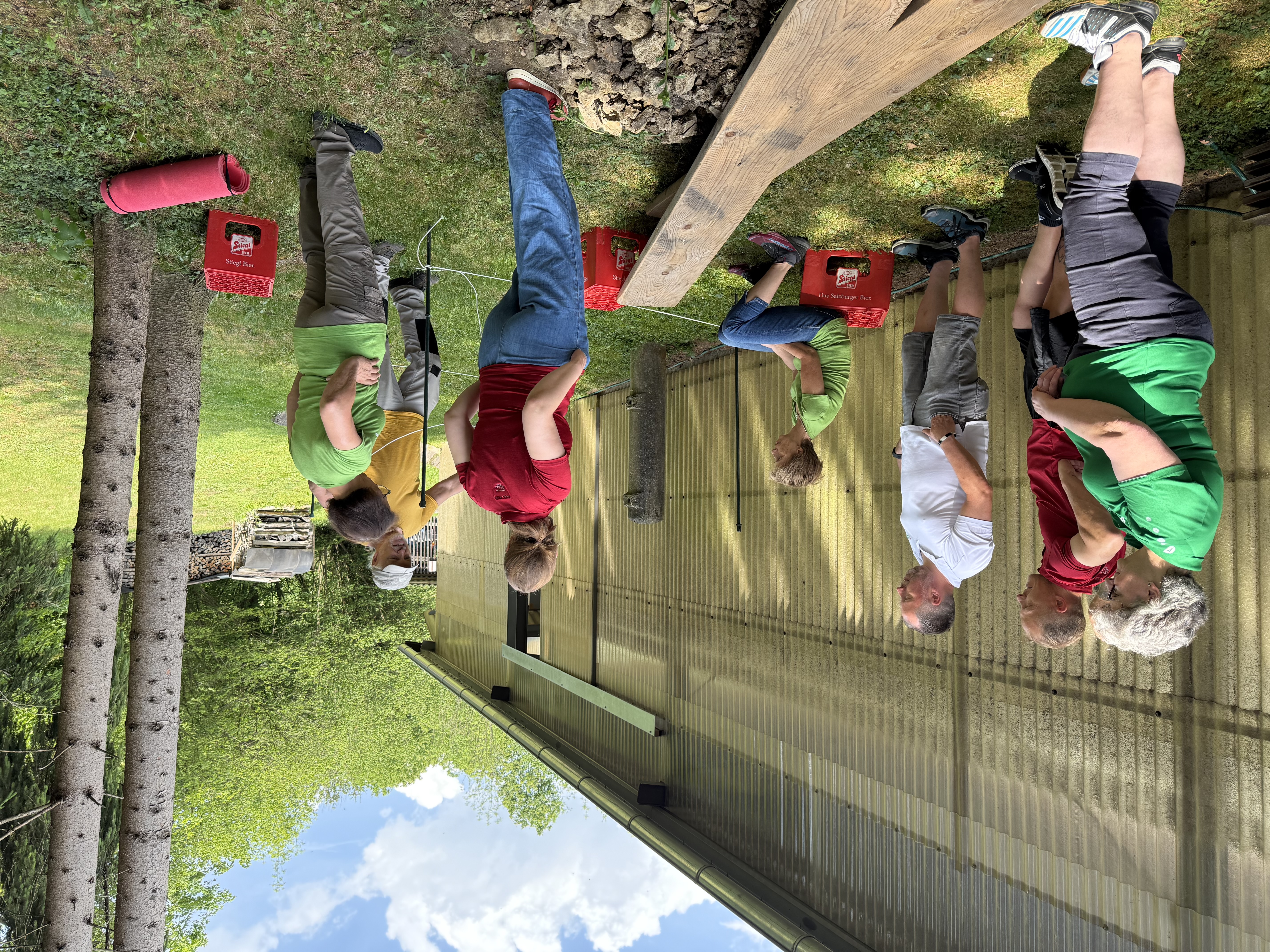 Konzentration und Teamwork: Die Gruppe beim praktischen Teil der Ausbildung im Freien. Gruppe von Personen steht im Freien vor einer Halle und beobachtet eine Erklärung zum Balancier-Übungsaufbau mit Holzbrettern und Bierkisten.